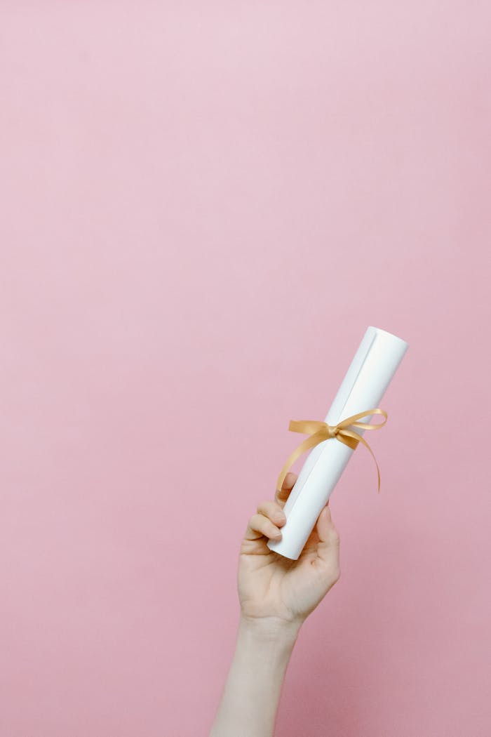 A hand holding a rolled diploma tied with a ribbon against a pink background.