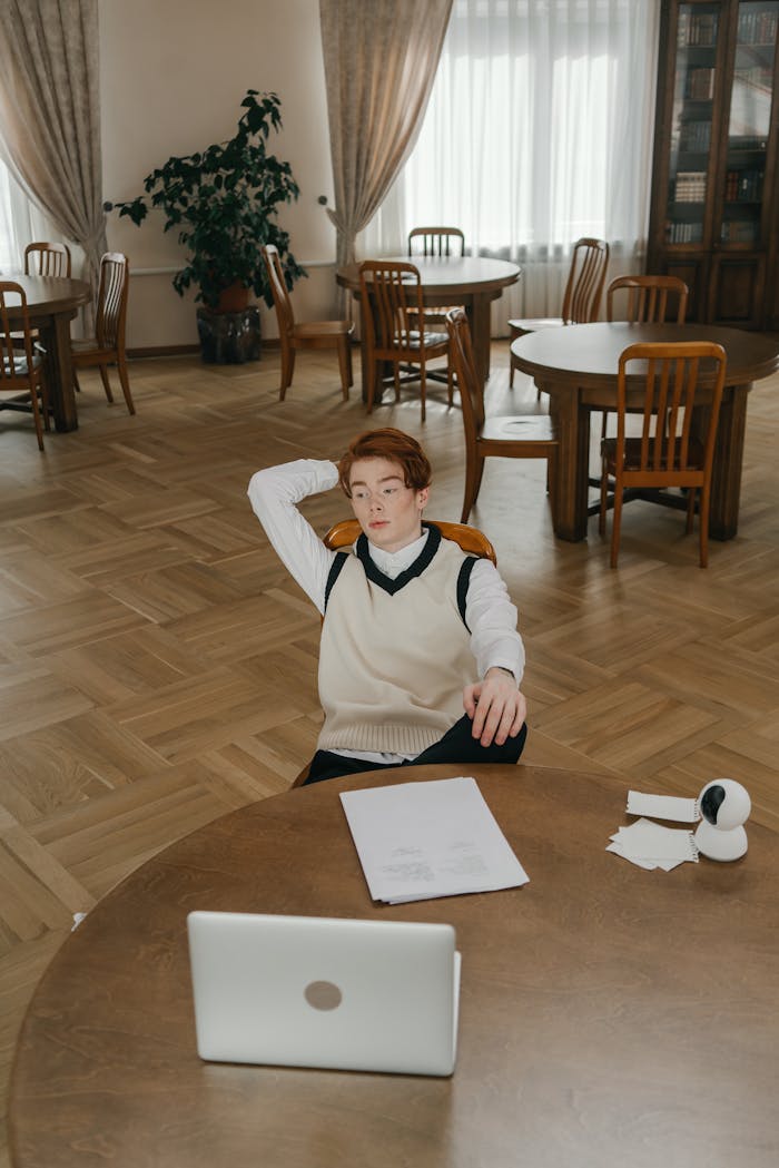 Young man in sweater vest sitting relaxed with laptop in a modern library interior.